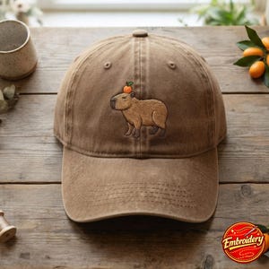 Puede incluir: Una gorra de béisbol marrón con un diseño bordado de un capibara beige de perfil con una pequeña fruta naranja sobre su cabeza. La gorra tiene una corona estructurada, una visera curva y detalles de costuras visibles en toda la tela.