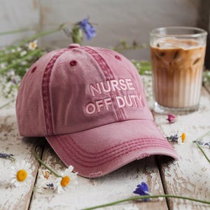 Puede incluir: Una gorra de béisbol rosa con el texto "NURSE OFF DUTY" bordado en blanco. La gorra tiene un aspecto desgastado y está hecha de una tela suave y cómoda.