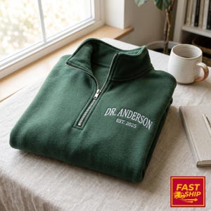 May include: A folded, forest green quarter-zip sweatshirt with white embroidered text that reads "DR. ANDERSON EST. 2025". The sweatshirt is made of a soft material and has a silver zipper. The item is displayed on a light-colored surface.