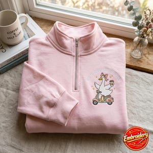 May include: A light pink quarter-zip sweatshirt featuring an embroidered graphic of a white goose wearing a pink bow, riding a scooter, and holding a coffee cup. The text above the goose reads "OFF TO REGULATE EMOTIONS" in white lettering.