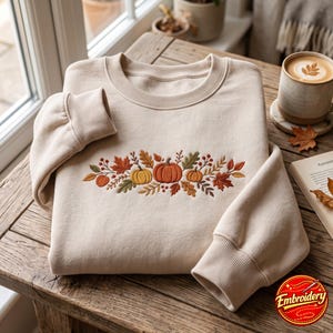 Könnte beinhalten: Ein beigefarbenes Sweatshirt mit Rundhalsausschnitt und einer zentralen Stickerei aus drei Kürbissen, umgeben von Herbstblättern, Beeren und Laub in Orange-, Gelb-, Braun- und Grüntönen. Das Kleidungsstück hat lange Ärmel mit gerippten Bündchen.