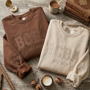 May include: Two crewneck sweatshirts, one brown and one beige, embroidered with "BCBA EST. 2026". The brown sweatshirt has "Kelsey" embroidered on the sleeve. The beige sweatshirt has "Liam" embroidered on the sleeve. A candle, walnuts, and books are in the background.