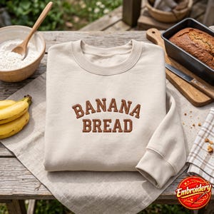 Puede incluir: Una sudadera de cuello redondo en color beige con el texto BANANA BREAD bordado en letras mayúsculas marrones en el pecho. La prenda tiene mangas largas con puños acanalados y un cuello acanalado, presentada sobre una superficie de tela clara.