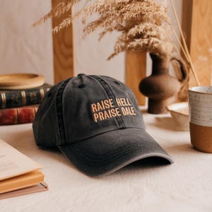 May include: A dark gray baseball cap with the embroidered text "RAISE HELL PRAISE DALE" in orange. The cap has a curved brim and a distressed look. The hat is sitting on a white surface.