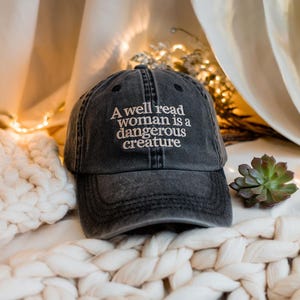 Puede incluir: Una gorra de béisbol negra desgastada con el texto "A well read woman is a dangerous creature" bordado en blanco.
