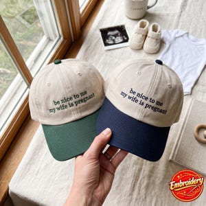 Puede incluir: Dos gorras de béisbol con coronas beige y viseras oscuras. La gorra izquierda tiene una visera verde oscuro y la derecha una visera azul marino. Ambas presentan el texto negro bordado: be nice to me my wife is pregnant, en el panel frontal.