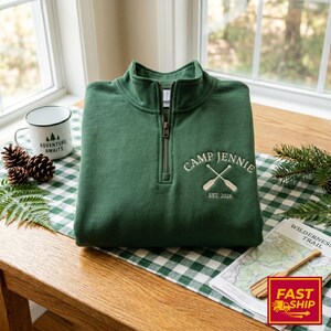 Könnte beinhalten: Ein waldgrüner Quarter-Zip-Pullover mit einer Stickerei auf der Brust, die den Text "CAMP JENNIE" über zwei gekreuzten Rudern und darunter "EST. 2026" zeigt. Das Kleidungsstück hat einen hohen Kragen mit einem Metallreißverschluss.