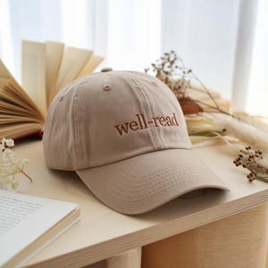May include: A tan baseball cap with the embroidered text "well-read" in a darker brown. The cap is made of a soft fabric and has a curved brim. The cap is sitting on a wooden surface.