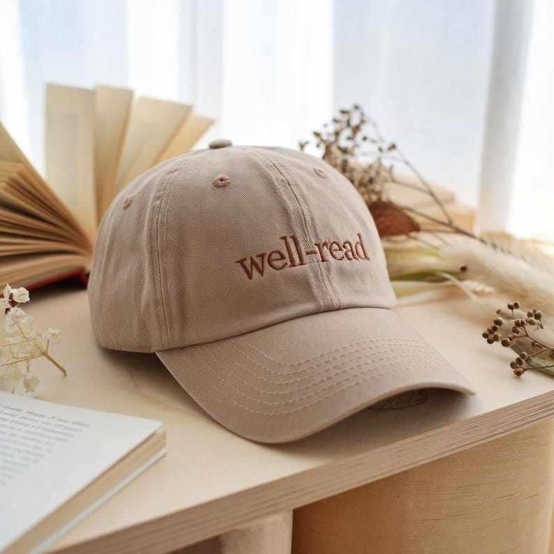 May include: A tan baseball cap with the embroidered text "well-read" in a darker brown. The cap is made of a soft fabric and has a curved brim. The cap is sitting on a wooden surface.