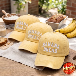 May include: Three yellow baseball caps with a distressed finish, each featuring the words BANANA BREAD embroidered in white capital letters on the front panel. The caps have a structured crown, a curved brim, and a button on the top center.