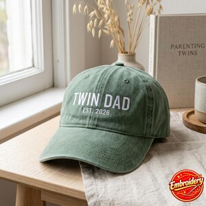 May include: A sage green baseball cap featuring white embroidered text that reads TWIN DAD EST. 2026. The cap has a structured crown, a curved brim with visible stitching, and eyelets on the top panels for ventilation, presented in a clean style.