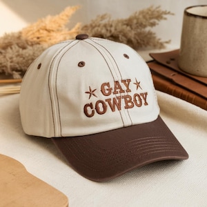 Peut inclure: Casquette de baseball avec une couronne crème et une visière marron. Le texte brodé "GAY COWBOY" en marron, avec des étoiles, orne la casquette. Les coutures et le bouton supérieur sont également marron.