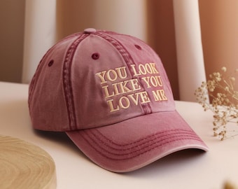 May include: A dusty rose baseball cap with the embroidered text "YOU LOOK LIKE YOU LOVE ME" in a gold color. The cap has a curved brim and a fabric strap closure.