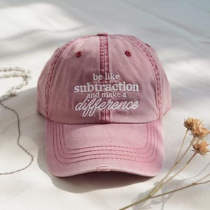 May include: A dusty rose baseball cap with the embroidered text "be like subtraction and make a difference" in white. The cap has a distressed look and a curved brim. The hat is a casual accessory.