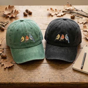 Könnte beinhalten: Zwei Baseballkappen, eine grüne und eine schwarze, liegen nebeneinander auf einer Holzoberfläche. Jede Kappe hat ein gesticktes Design von drei Vögeln in Gelb, Rot und Blau. Die Kappen haben einen klassischen, abgenutzten Look.