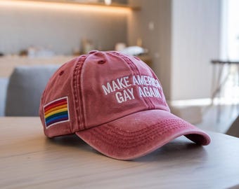 Può includere: Un berretto da baseball rosso con il testo "Make America Gay Again" ricamato in bianco. Una bandiera arcobaleno del pride è cucita sulla parte anteriore del berretto.