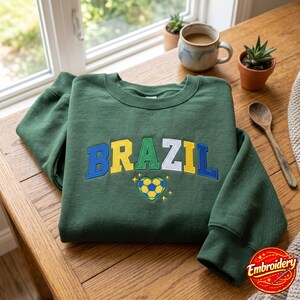 Peut inclure: Un sweat-shirt à col rond vert foncé avec le mot BRAZIL brodé en lettres capitales bleues, jaunes, vertes et blanches sur la poitrine. Sous le texte se trouve un motif de ballon de football en forme de cœur avec des étoiles jaunes.