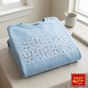 Könnte beinhalten: Hellblaues Sweatshirt mit dem gestickten Schriftzug "SOCIAL WORKER" in Weiß mit floralem Design. Das Sweatshirt ist ordentlich auf einem Tisch gefaltet. Eine Tasse und ein Buch sind im Hintergrund zu sehen.