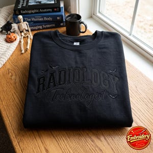 Puede incluir: Una sudadera negra de cuello redondo con bordado negro tonal que dice RADIOLOGY Technologist en una fuente universitaria, acentuada con telarañas y pequeños murciélagos bordados que rodean el texto en la parte frontal.