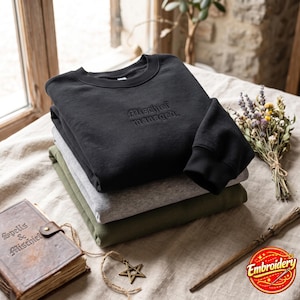 May include: A stack of three crewneck sweatshirts in black, heather grey, and olive green. The top black sweatshirt features the embroidered text "Mischief managed." in a dark thread. The garments have ribbed collars and sleeve cuffs.