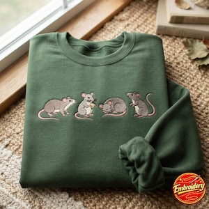 Könnte beinhalten: Ein waldgrüner Rundhals-Pullover mit einer Stickerei von vier kleinen grauen Mäusen in verschiedenen Posen. Die Mäuse sind in einer Reihe dargestellt, wobei eine steht, eine einen kleinen Gegenstand hält, eine zusammengerollt schläft und eine aufrecht steht.