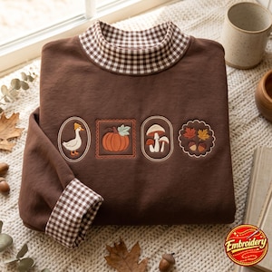 Op de afbeelding: Een bruin sweatshirt met lange mouwen, een opstaande kraag en mouwboorden met een bruin-wit vichy-ruitpatroon. De voorzijde is voorzien van vier borduursels: een gans met een muts, een pompoen, paddenstoelen en herfstbladeren met eikels.