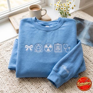 Peut inclure: Un sweat-shirt bleu clair à col rond avec des icônes médicales brodées en blanc, incluant un nœud, un scanner CT, un symbole de radiation, une radiographie thoracique et un stéthoscope. Le prénom Olivia est brodé en blanc sur la manche.
