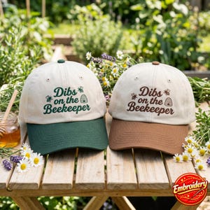 May include: Two baseball caps with off-white crowns and embroidered text reading Dibs on the Beekeeper. One cap features a dark green brim and green text, while the other has a brown brim and brown text. Both include small bee and beehive icons.
