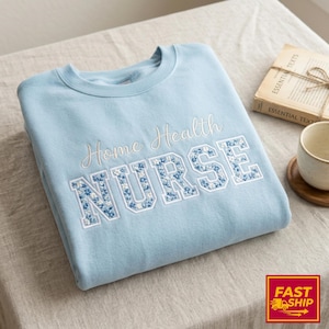 May include: A light blue sweatshirt with the embroidered text "Home Health NURSE." The word "NURSE" is in a block letter style with a floral pattern in shades of blue and white. A book and a cup are in the background.