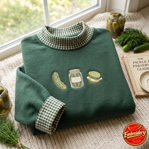 May include: A dark green sweatshirt featuring a mock neck and cuffs with a white and green gingham pattern. The chest is embroidered with a single pickle, a glass jar filled with pickles, and a sliced pickle sandwich on a light background.