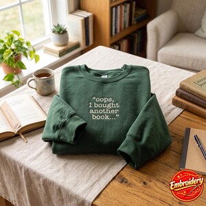 May include: A forest green crewneck sweatshirt featuring white embroidered text on the chest that reads, "oops, I bought another book..." The garment has long sleeves and a ribbed collar, cuffs, and hem, presented in a folded, flat-lay style.