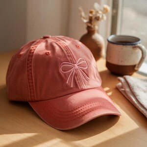 Puede incluir: Una gorra de béisbol rosa con un lazo blanco bordado en la parte delantera. La gorra tiene una visera curvada y una parte trasera de malla.