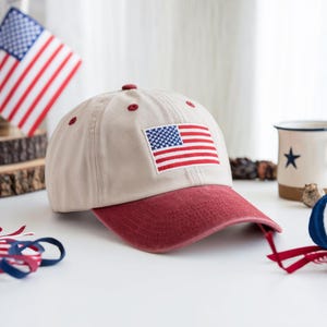 Puede incluir: Gorra de béisbol beige con visera roja y la bandera americana bordada en la parte delantera. La gorra tiene botones rojos en la parte superior. La imagen también incluye una pequeña bandera americana y una taza con una estrella.