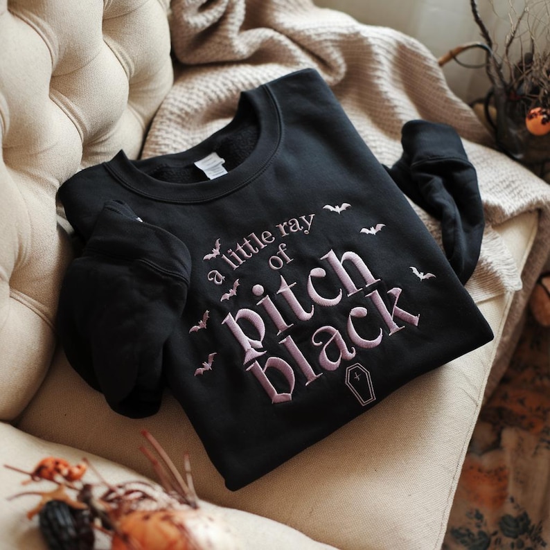 May include: Black sweatshirt featuring the embroidered phrase "a little ray of Pitch Black" in pink, with bat and coffin motifs. The garment is shown on a beige armchair.