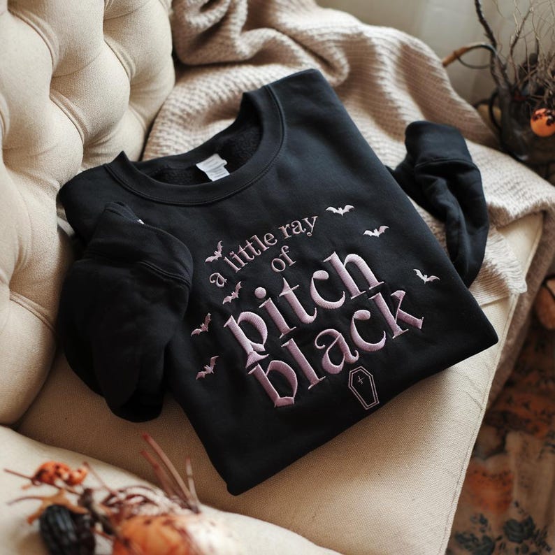 May include: Black sweatshirt featuring the embroidered phrase "a little ray of Pitch Black" in pink, with bat and coffin motifs. The garment is shown on a beige armchair.