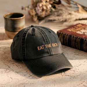 Könnte beinhalten: Eine schwarze Baseballkappe mit einem goldenen, gestickten Slogan, der "EAT THE RICH" lautet.