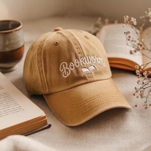 Op de afbeelding: Een beige baseballpet met een wit geborduurd ontwerp met de tekst "Bookworm" en een open boekicoon. De pet ligt op een wit oppervlak met een boek en bloemen.