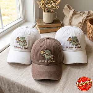 Puede incluir: Dos gorras de béisbol blancas y una gorra de béisbol marrón, cada una con una rana verde bordada que sostiene una lámpara y un cuadro, con el texto WE THRIFT AT DAWN bordado debajo del gráfico en la parte frontal de cada gorra.