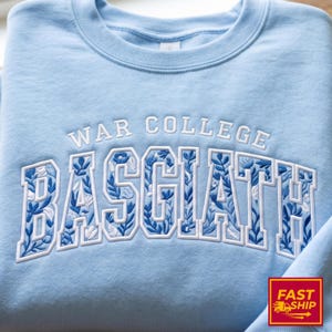 Puede incluir: Sudadera azul claro con las palabras "WAR COLLEGE BASCIATH" bordadas en blanco. Las letras de "BASCIATH" est&aacute;n rellenas con un dise&ntilde;o floral azul. La sudadera tiene cuello redondo.