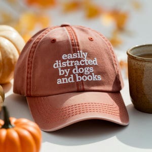 Op de afbeelding: Een roze baseballpet met de geborduurde tekst "easily distracted by dogs and books" in het wit. De pet ligt op een wit oppervlak met een pompoen en een mok thee op de achtergrond.