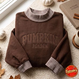 May include: A dark brown sweatshirt featuring a mock neck collar and sleeve cuffs in a brown and white gingham pattern. The front of the garment displays the embroidered text PUMPKIN SEASON in a dark brown arched font across the chest area.