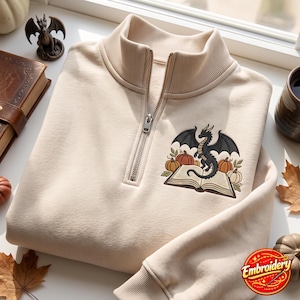 Op de afbeelding: Een beige sweater met een korte rits en een geborduurd ontwerp op de borst. De afbeelding toont een zwarte draak op een open boek, omringd door oranje en crèmekleurige pompoenen en groene bladeren. De sweater heeft een geribbelde kraag.