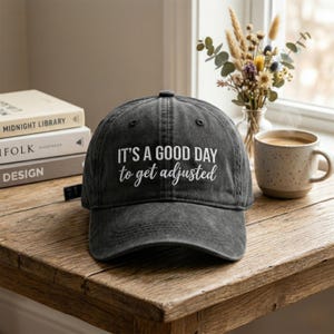 Peut inclure: Casquette de baseball gris foncé avec l'inscription blanche "IT'S A GOOD DAY to get adjusted". La casquette est posée sur une table en bois, avec des livres et une tasse de café en arrière-plan.