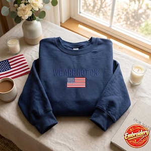 Può includere: Una felpa girocollo blu navy con il testo ricamato "WE ARE NOT OK" in filo blu abbinato, posizionato sopra una piccola e dettagliata bandiera americana ricamata con cuciture rosse, bianche e blu sul petto.