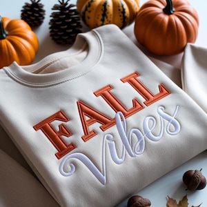 Peut inclure: Sweat-shirt crème avec le texte brodé "FALL Vibes" en orange et blanc. L'image comprend des citrouilles, des pommes de pin et des glands, suggérant un thème automnal. Le sweat-shirt est de style col rond.