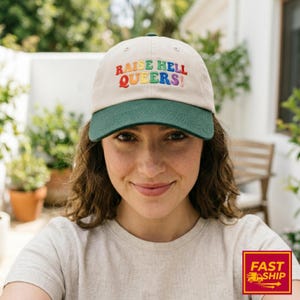May include: A beige baseball cap with a dark green brim. The front of the cap features the embroidered text "RAISE HELL QUEERS!" in a multi-colored rainbow gradient font. The cap has a structured crown with small ventilation eyelets at the top.