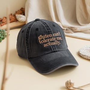Peut inclure: Casquette de baseball noire délavée avec la phrase brodée "gluten can't tolerate me, actually." Le chapeau a une visière incurvée et un bouton sur le dessus. Le texte est de couleur marron clair.