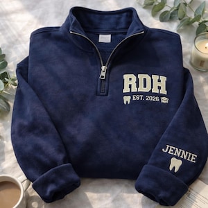 以下が含まれることがあります： 刺繍とグラフィックが施されたネイビーブルーのクォータージップスウェットシャツ。「RDH EST. 2026」の文字と歯のグラフィックが前面にあります。袖には「JENNIE」の名前と歯のグラフィック。カジュアルで快適なトップス。