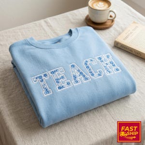 Könnte beinhalten: Hellblaues Sweatshirt mit dem Wort "TEACH", das in Weiß und Blau mit floralem Muster gestickt ist. Das Sweatshirt ist ordentlich auf einer neutralfarbenen Oberfläche gefaltet. Eine Kaffeetasse und ein Buch sind im Hintergrund.