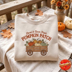 Pode incluir: Uma camisola de gola redonda cor de creme com um gráfico bordado de uma carroça de madeira cheia de abóboras. O texto acima da carroça diz Local & Farm Fresh PUMPKIN PATCH em letras cor de laranja e castanhas, com pequenos detalhes florais.
